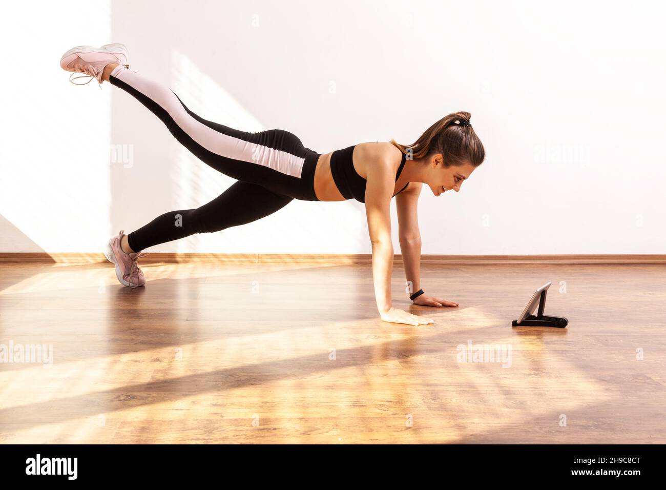 Mujer haciendo plancha con la pierna elevada, viendo vídeo tutorial en la  tableta, repetir después de blogger fitness, con ropa de deporte negro y  medias. Estudio completo, iluminado por la luz del