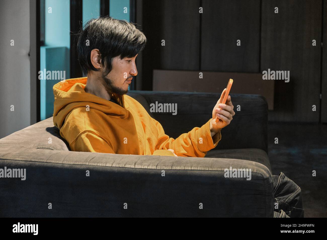 Un tipo asiático se sienta en un sofá en una pequeña oficina y se comunica por teléfono móvil. El concepto de la pequeña empresa y la comunicación en línea. Foto de stock