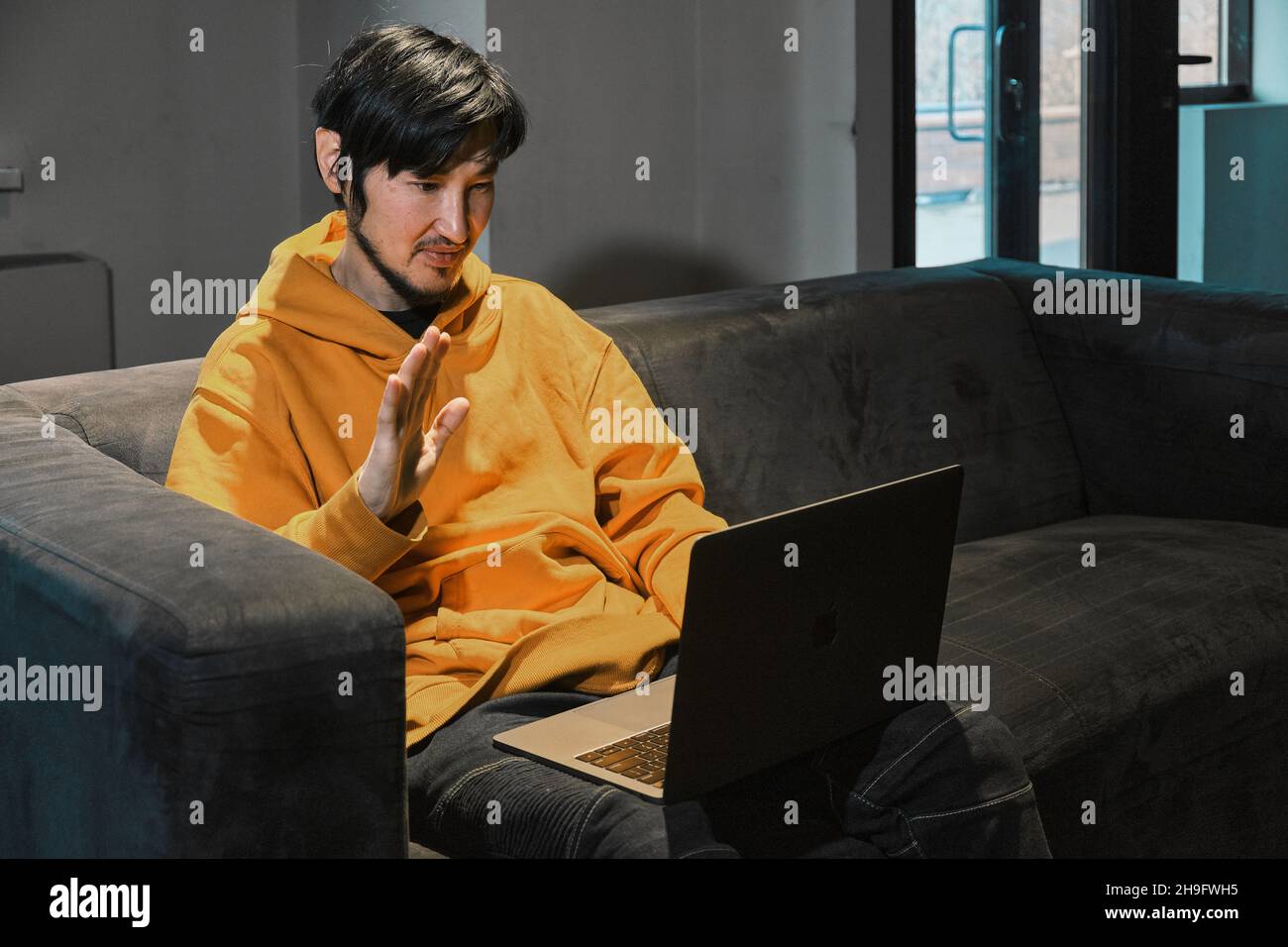 Un tipo asiático se sienta en un sofá en una pequeña oficina y se comunica a través de un enlace de vídeo a través de un ordenador portátil. El concepto de la pequeña empresa y la comunicación en línea. Foto de stock