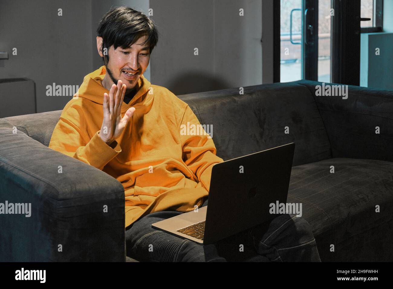 Un tipo asiático se sienta en un sofá en una pequeña oficina y se comunica a través de un enlace de vídeo a través de un ordenador portátil. El concepto de la pequeña empresa y la comunicación en línea. Foto de stock