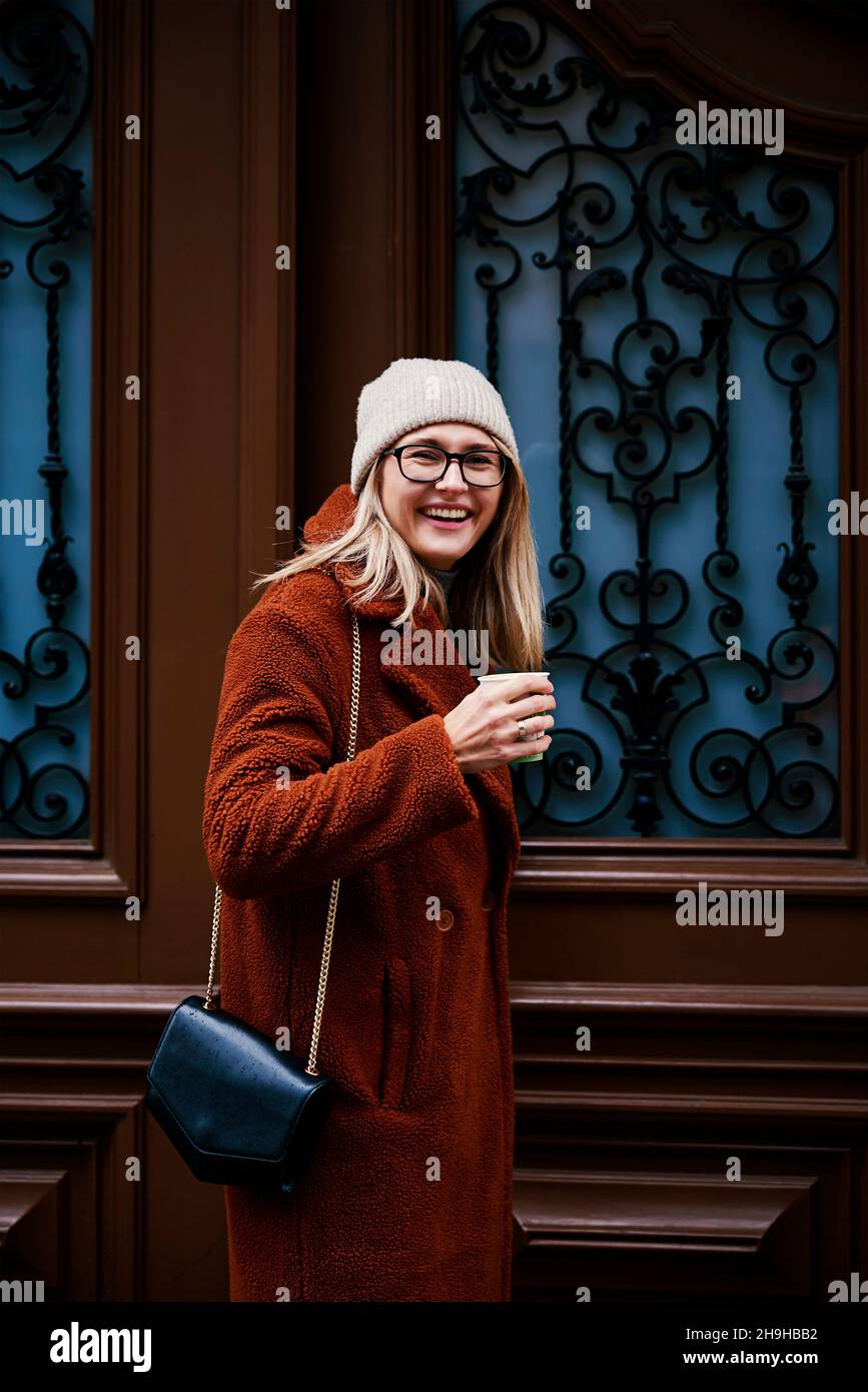 Mujer con abrigo y gafas con taza de café sonriendo y entrada en la puerta. Retrato de emoción feliz Foto de stock