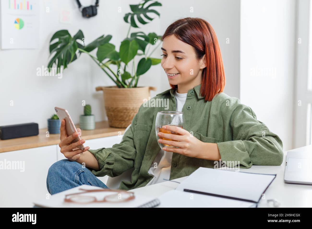 Mujer usando el teléfono móvil y bebiendo té. Joven empresaria en un lugar de trabajo cómodo. Concepto de negocio o comunicación en línea, oficina en casa y. Foto de stock