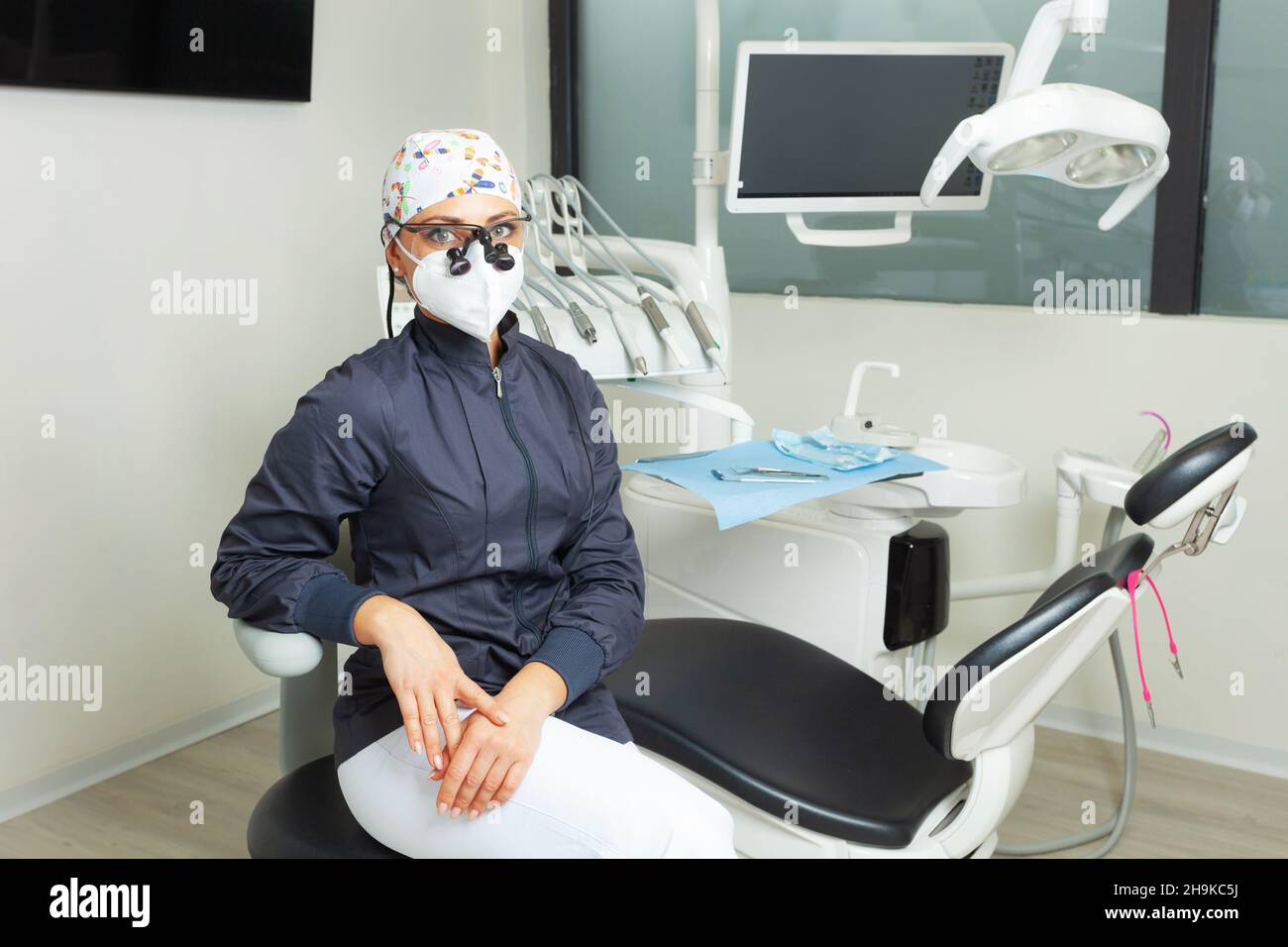 Hermosa mujer dentista sentada cerca de la silla dental. Retrato con equipo de protección personal y gafas binoculares. Foto de stock