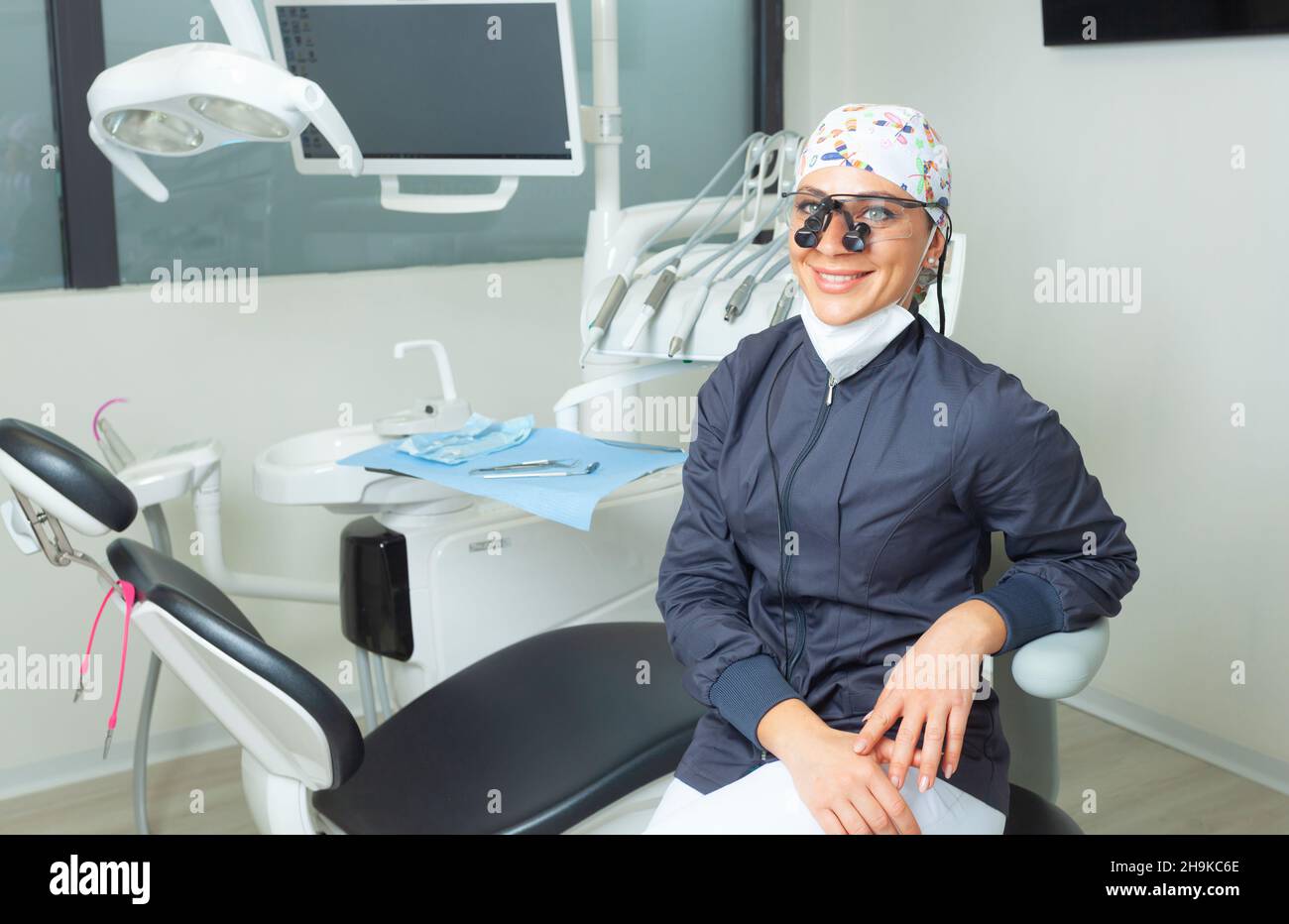 Hermosa mujer dentista sentada cerca de la silla dental. Retrato con equipo de protección personal y gafas binoculares. Foto de stock