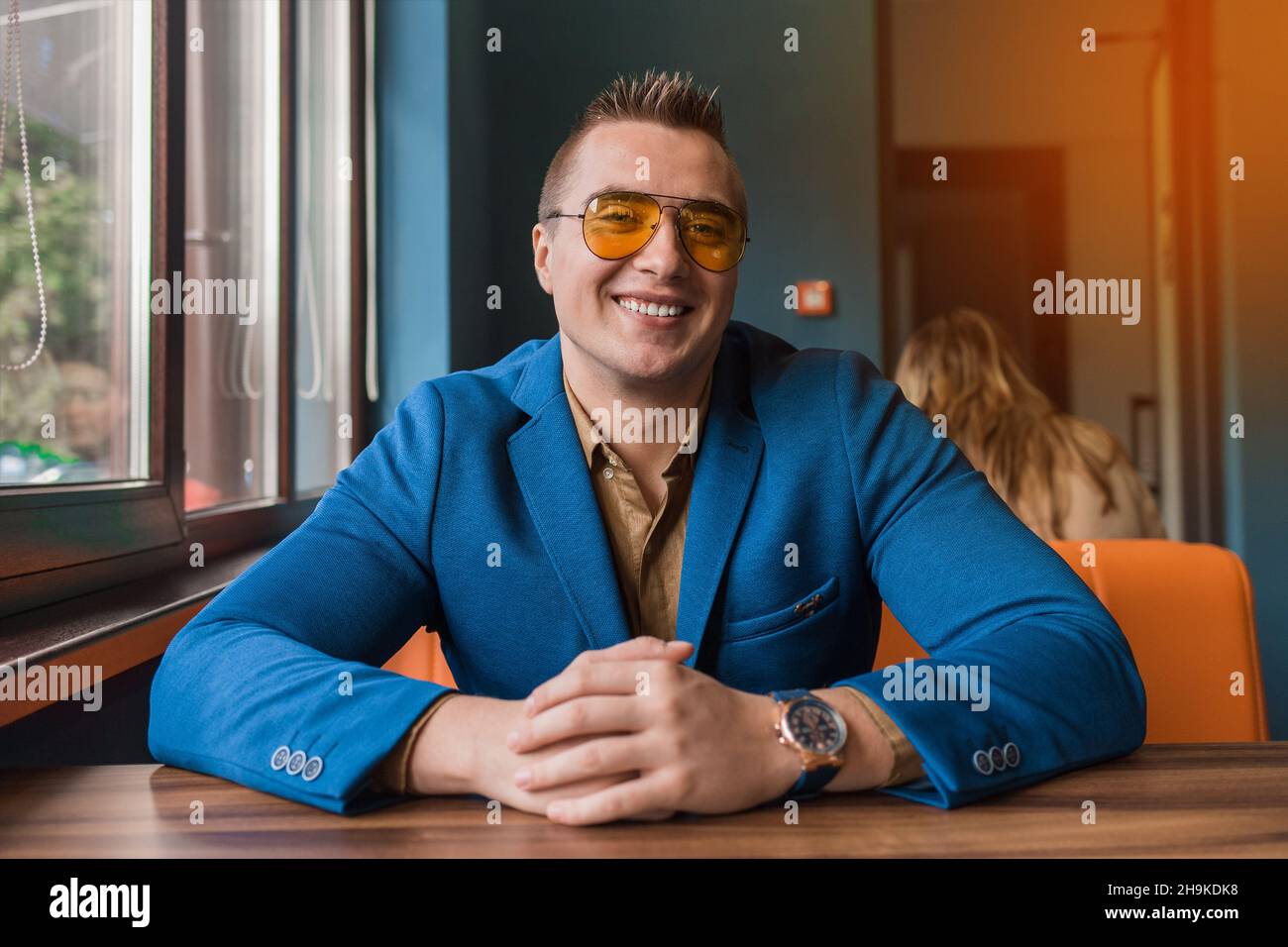 Hombre de negocios sonriente lindo positivo de aspecto caucásico retrato con estilo en gafas de sol, chaqueta y camisa, sentado en una mesa en un café de dos por dos. Foto de stock