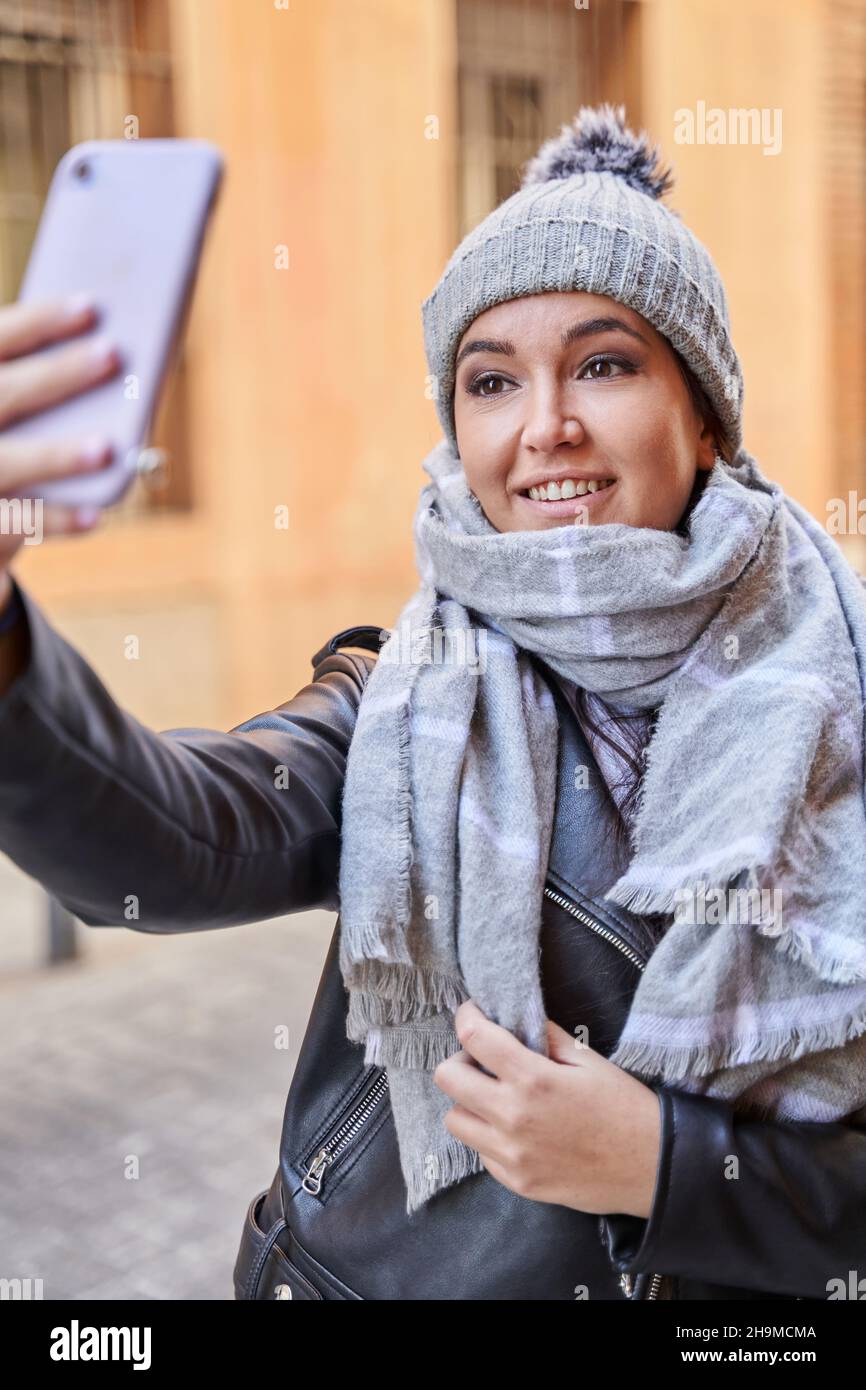 Mujer joven con estilo en ropa exterior sonriendo y tomando selfie mientras que parada en la calle de la ciudad en día frío Foto de stock