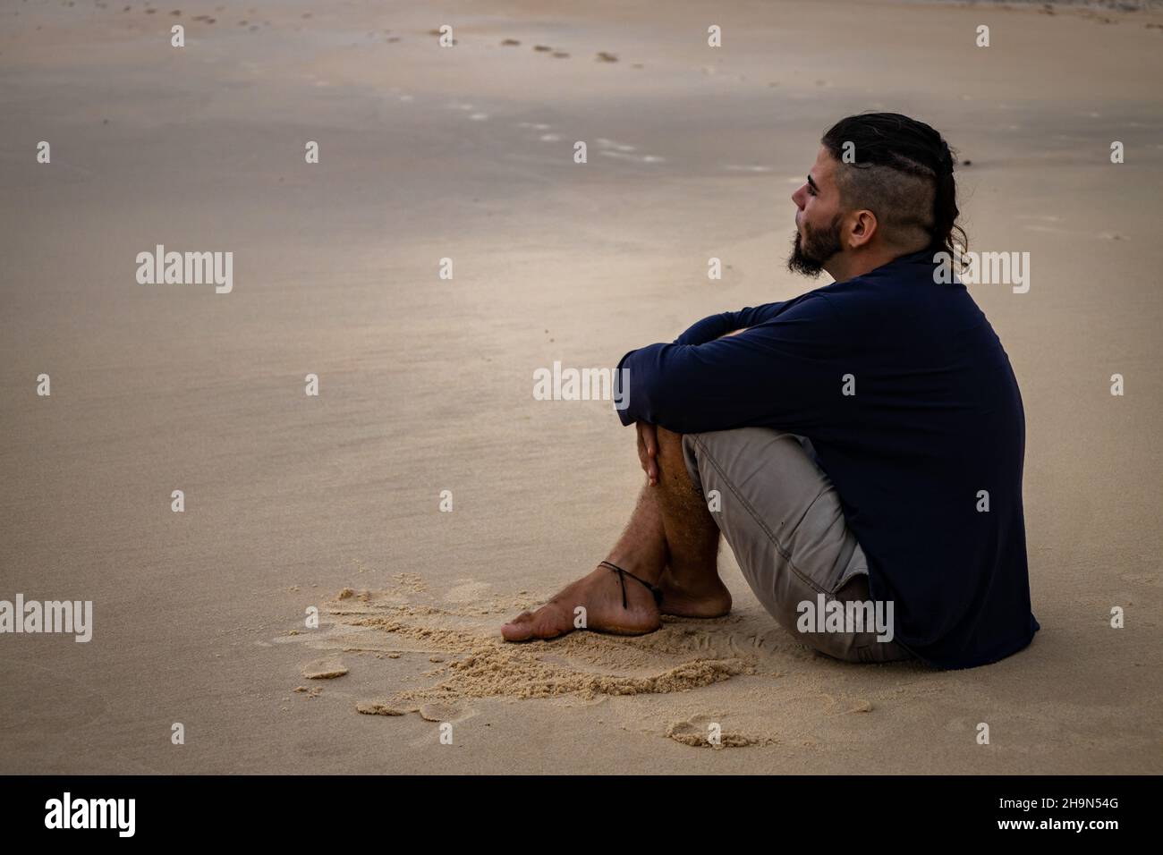Hombre hispano de 25 a 30 años y aspecto hippie sentado en una playa con sus piernas cruzadas. Espacio libre para escribir. Concepto de depresión o lonelina Foto de stock