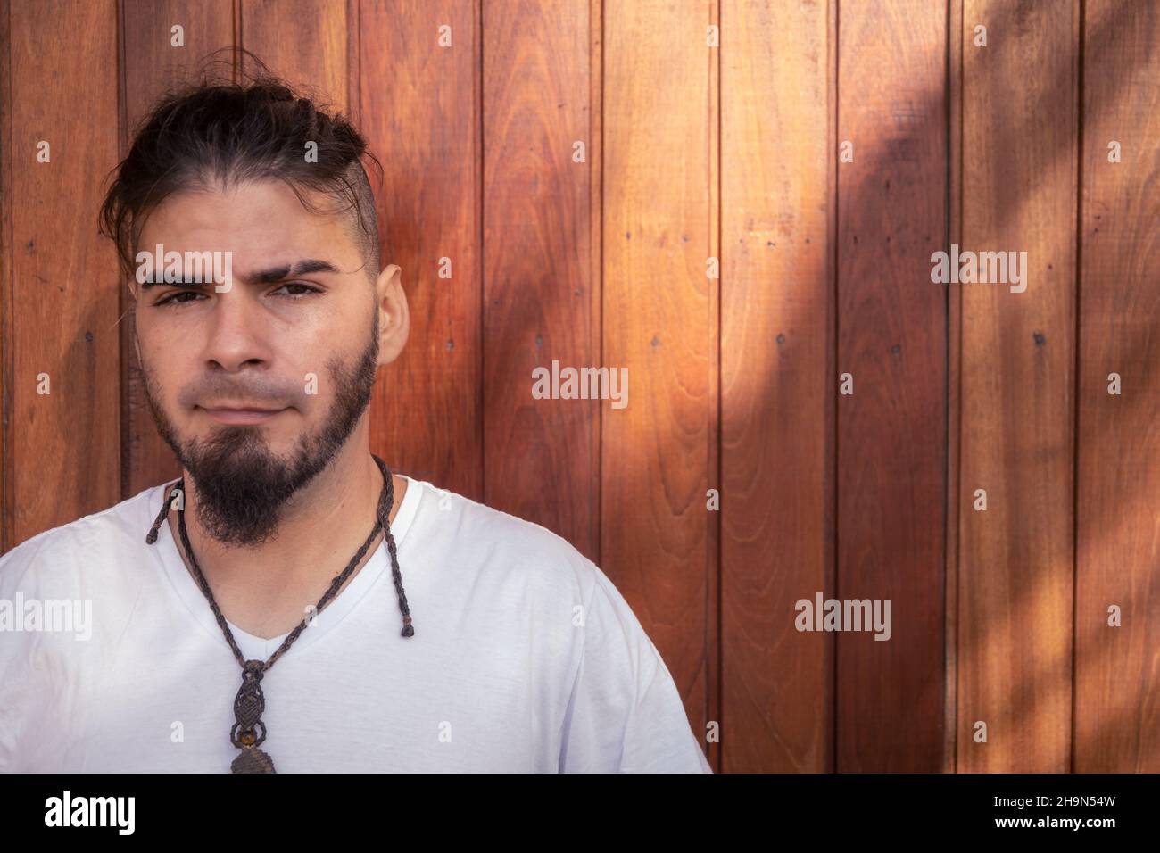 Hombre de 25 a 30 años de raza hispana y aspecto hippie mirando directamente a la cámara con un fondo de madera gastado. Foto de stock