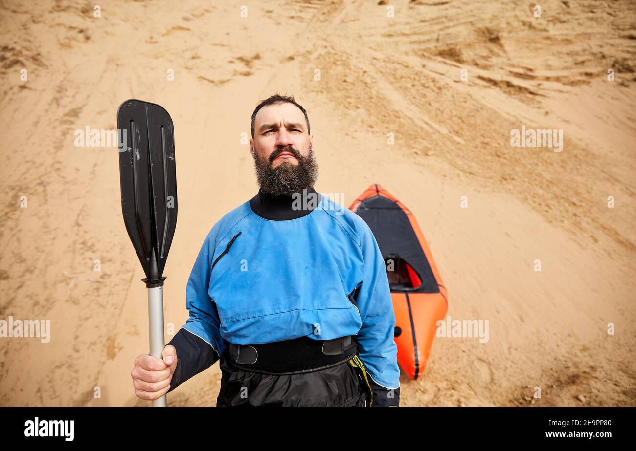 Retrato del anciano con barba negra y remar en chaqueta azul cerca de bote deportivo naranja balsa en fondo de dunas de arena Foto de stock