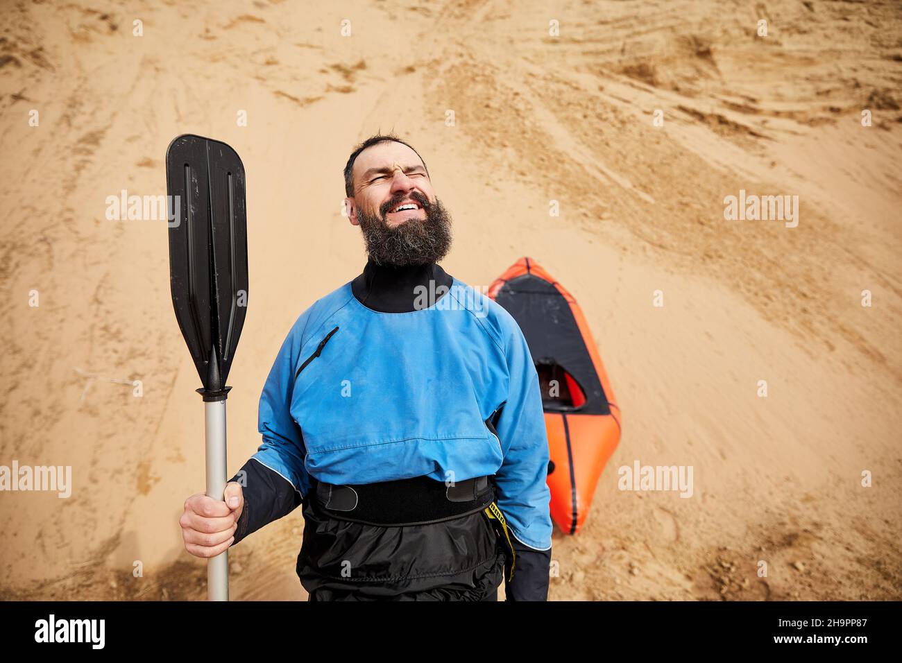 Retrato del anciano con barba negra y remar en chaqueta azul sonriendo cerca de bote deportivo naranja en fondo de dunas de arena Foto de stock