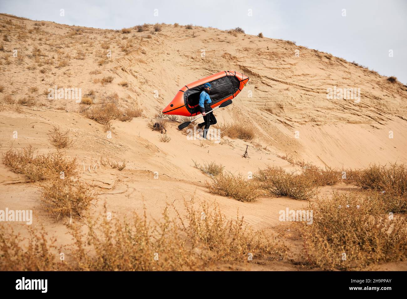 Anciano con barba negra en chaqueta azul sosteniendo bote deportivo naranja balsa en dunas de arena en el fondo del desierto. Concepto de aventura y viaje. Foto de stock