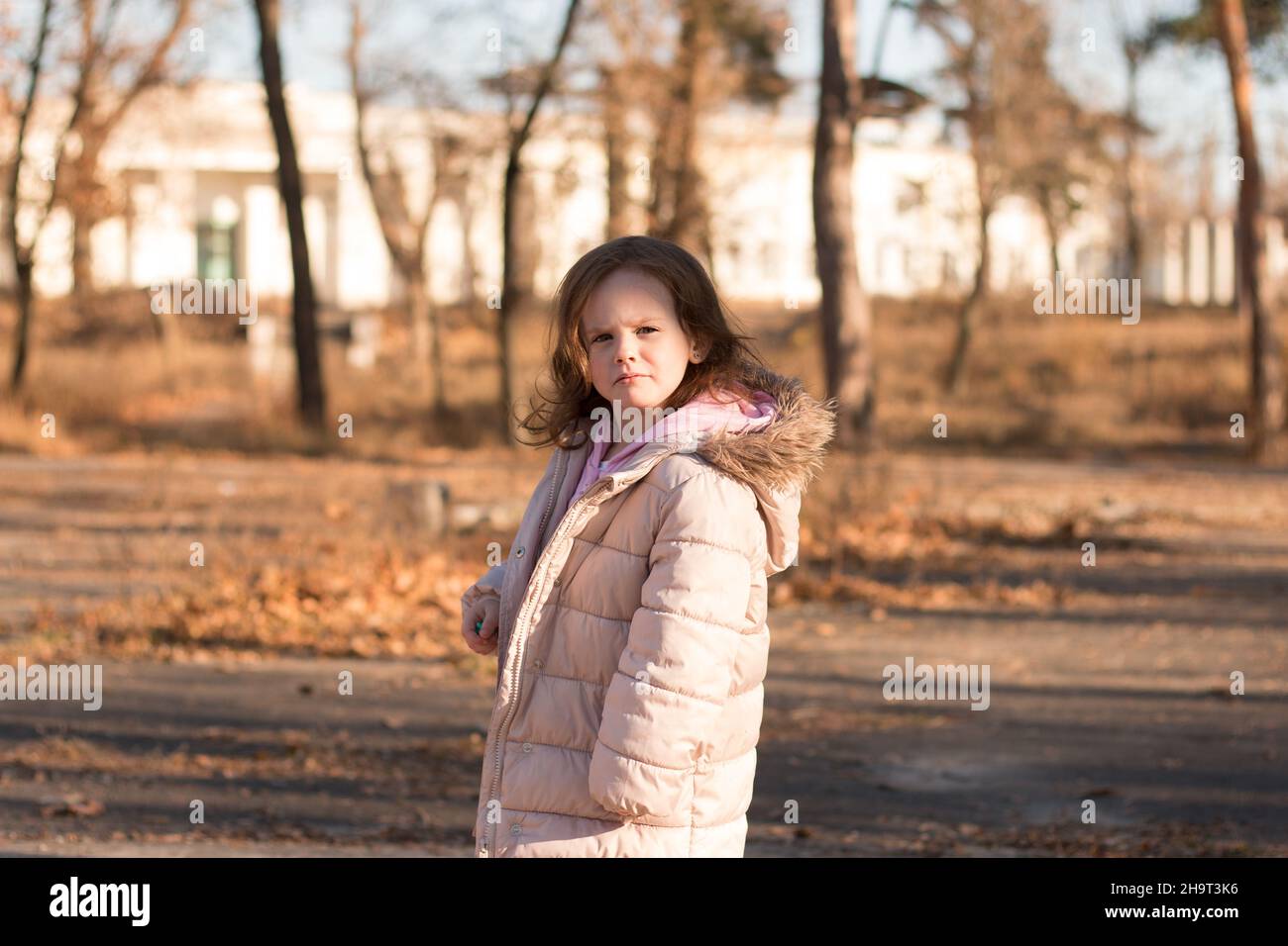 Una chica solitaria en una chaqueta una capucha esponjosa da la y mira a la cámara. Niño con ropa de abrigo en una calle fría de otoño Fotografía de