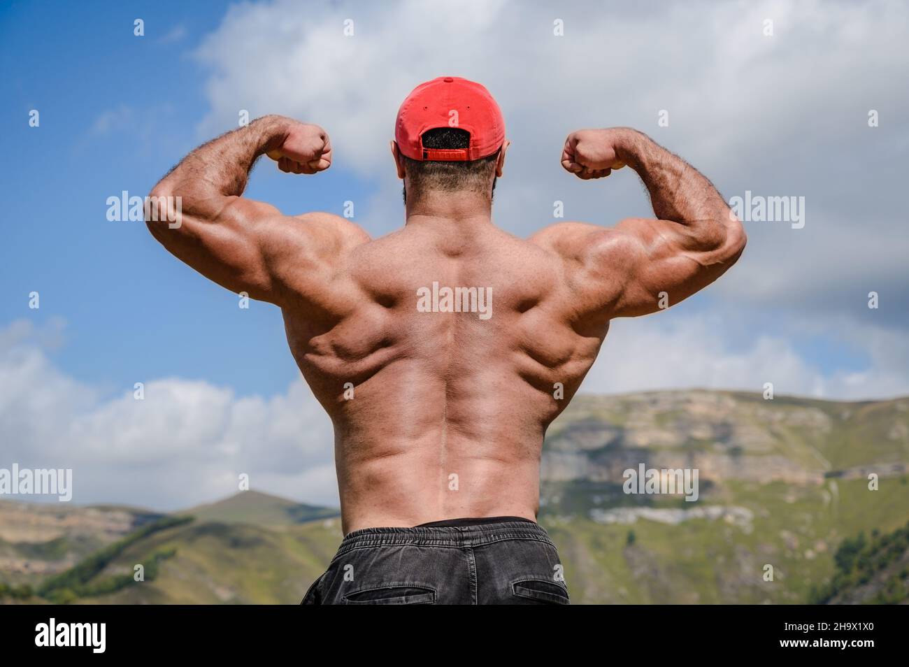 varón joven fuerte con músculos de la espalda grandes mostrando bíceps dobles sobre fondo alto de la montaña Foto de stock