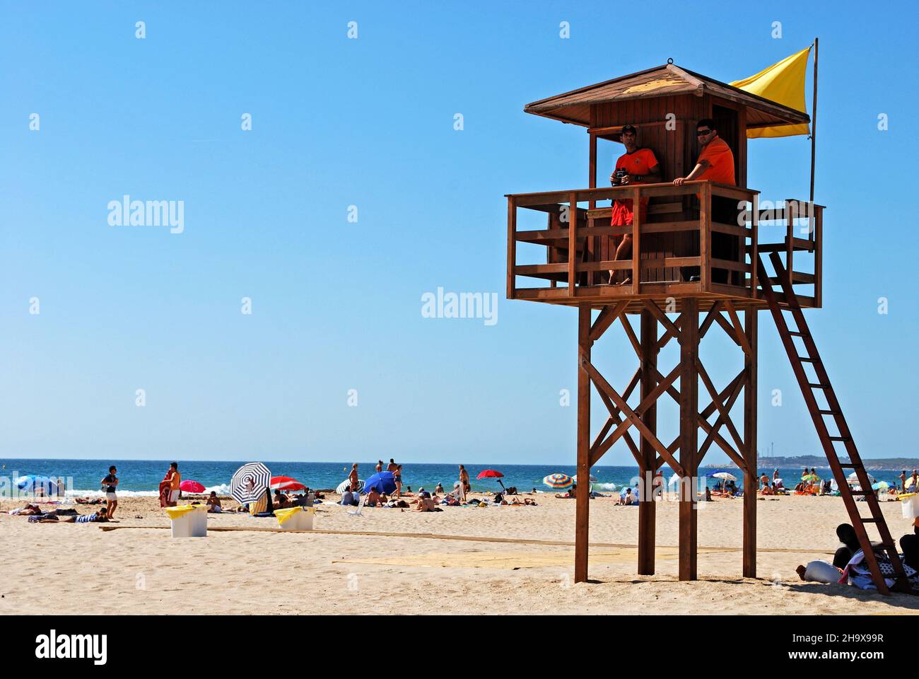Socorristas en una torre de madera en la playa con turistas disfrutando del entorno, Conil de la Frontera, España. Foto de stock