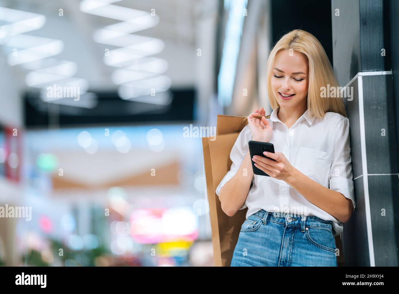 Vista en ángulo bajo de una mujer joven rubia sonriente y atractiva con ropa elegante utilizando el teléfono móvil sosteniendo bolsas de papel con compras. Foto de stock