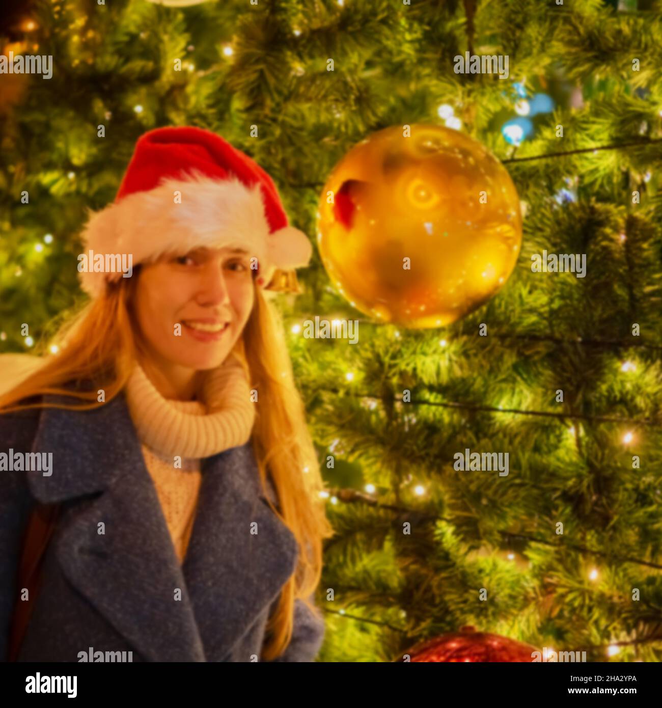 Imagen borrosa de una chica en sombrero de Santa cerca de un árbol de Navidad con grandes bolas. Imagen desenfoque Foto de stock