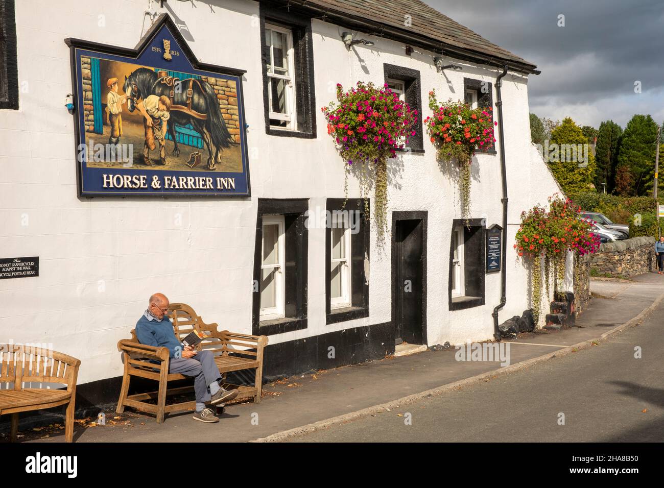 Reino Unido, Cumbria, Allerdale, Keswick, Threlkeld, Un hombre leyendo bajo el sol bajo el cartel del pub Horse and Farrier Foto de stock