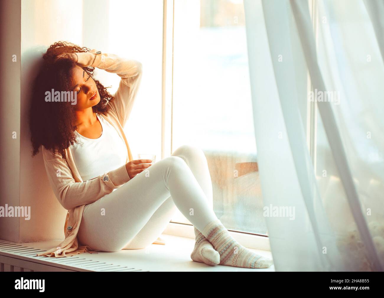 Feliz relajado africano americano etnia chica en el hogar sentado en el alféizar de la ventana contra el telón de fondo de cortinas revoloteando de viento ligero, feliz afro Foto de stock