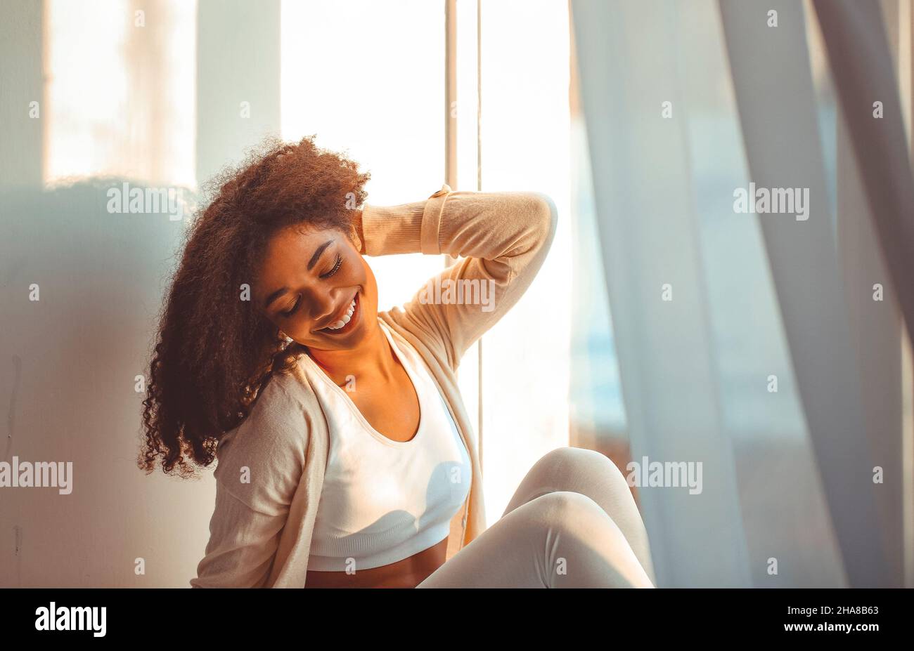 Feliz relajado africano americano etnia chica en el hogar sentado en el alféizar de la ventana contra el telón de fondo de cortinas revoloteando de viento ligero, feliz afro Foto de stock