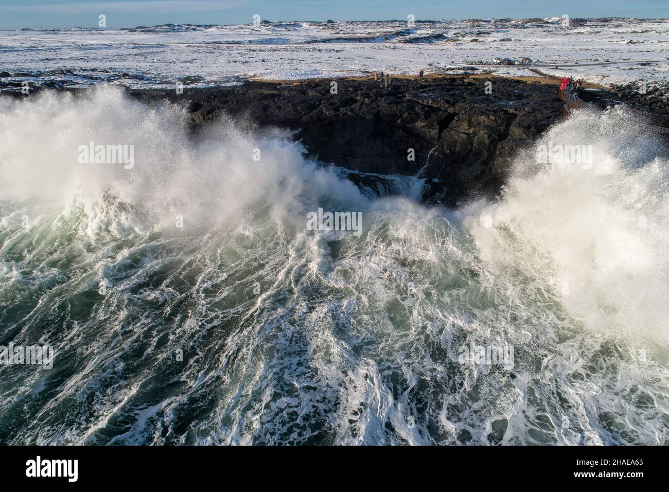 Disparo aéreo de las enormes olas del océano rompiendo en una orilla rocosa, grupo de turistas mirándolos muy pequeños en comparación. Foto de stock