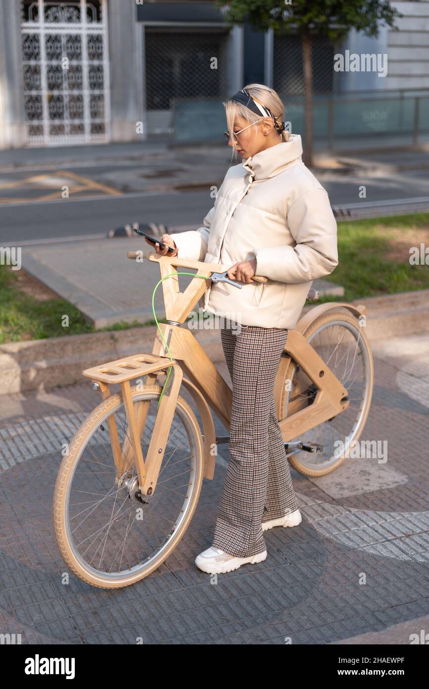 Alto ángulo de mujer adulta con ropa exterior elegante con smartphone mientras se encuentra en el pavimento cerca de una bicicleta ecológica de madera. Foto de stock