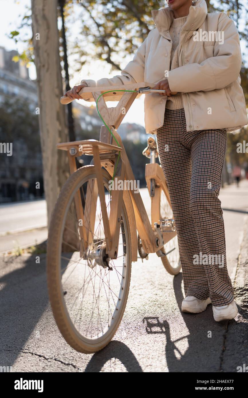 Mujer irreconocible en ropa exterior de pie cerca de una bicicleta ecológica de madera en un día soleado en la calle de la ciudad. Foto de stock