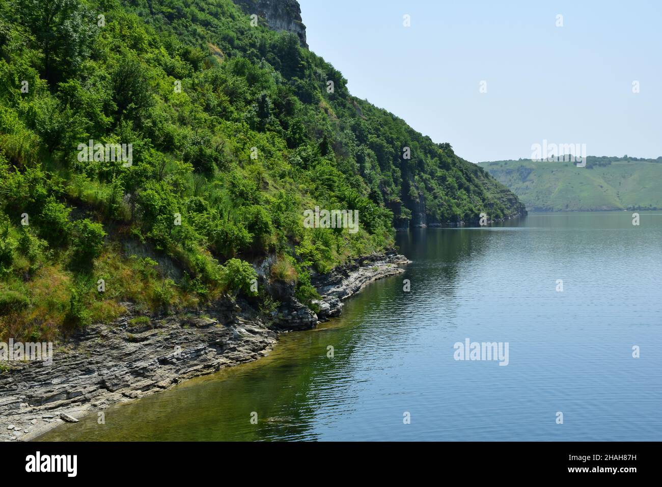 La orilla rocosa del lago está sobrecultivada con árboles verdes y arbustos al pie de la montaña Foto de stock