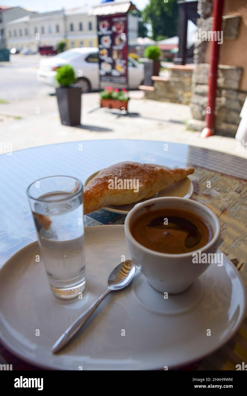 Una taza de café, un cruasán y un vaso de agua en un platillo están sobre una mesa en un café al borde de la carretera. El fondo está muy borroso Foto de stock