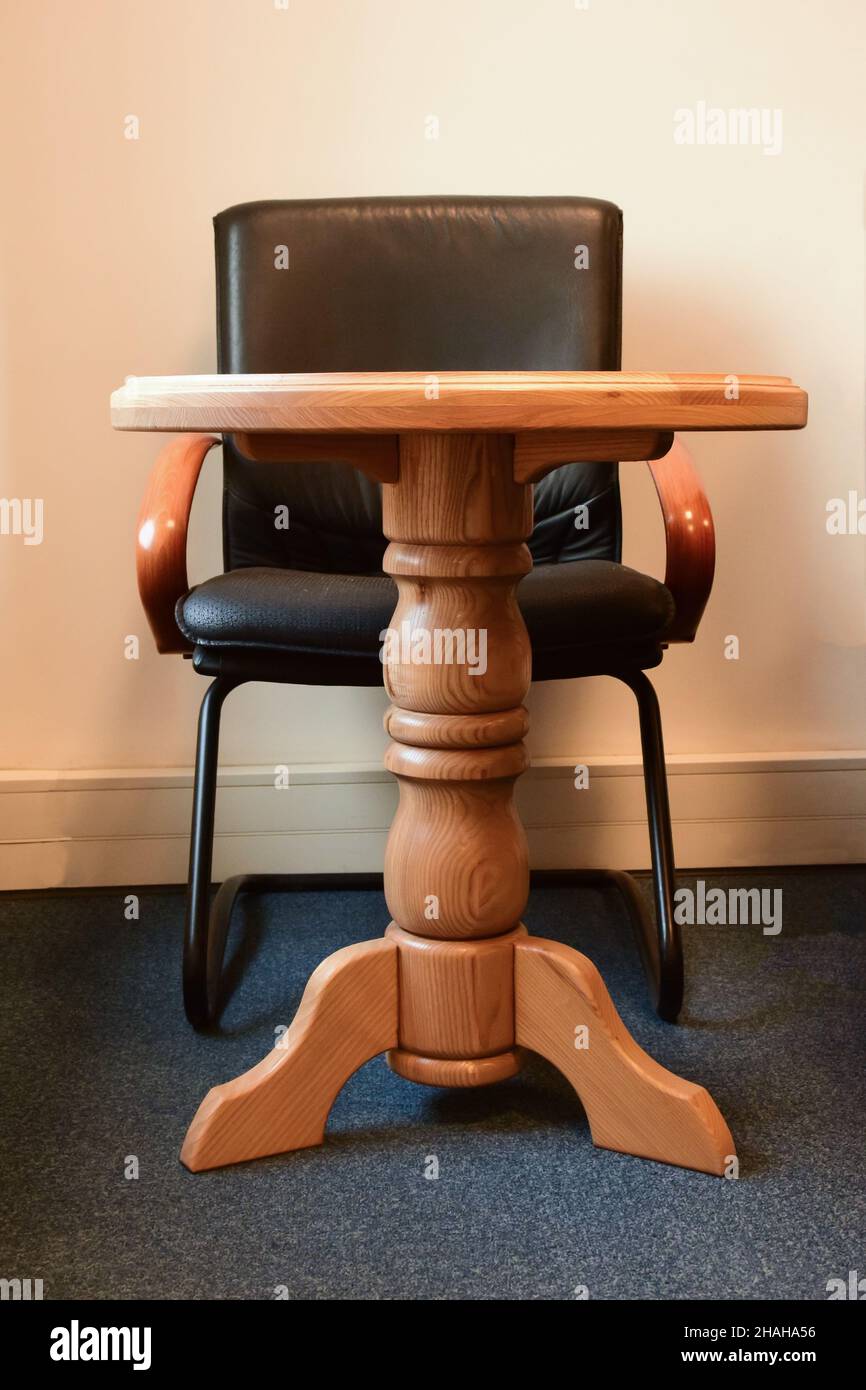 Decorativa mesa de madera sobre una pierna y detrás de ella un sillón de cuero negro sobre el fondo de la pared de la oficina Foto de stock
