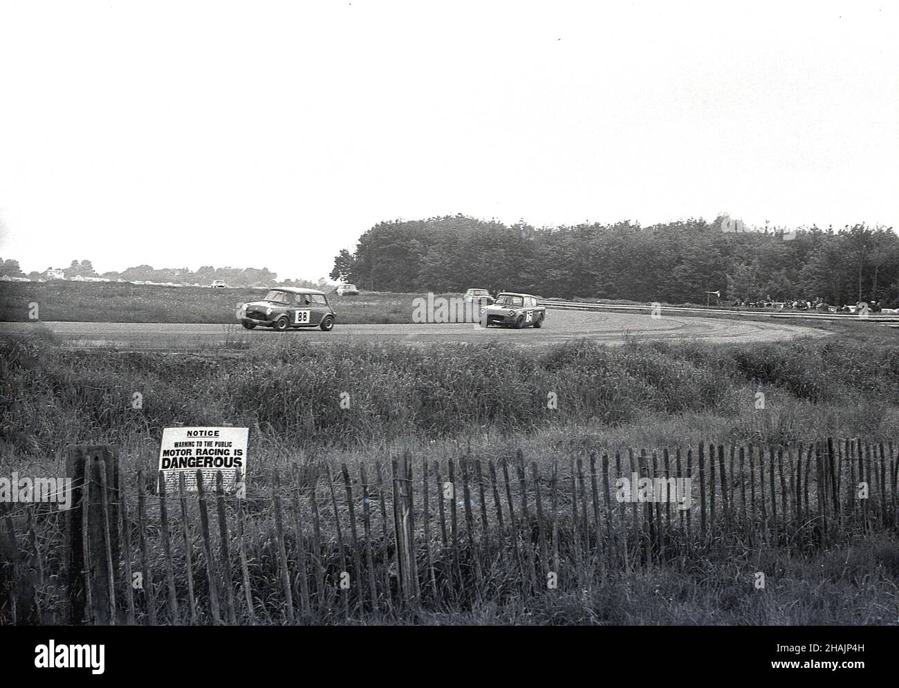 1960s, histórico, deporte de motor, carreras de coches, Inglaterra, REINO UNIDO. Lejos de la pista, un pequeño aviso en la hierba por una cerca de madera corta dice, Motor Racing es PELIGROSO. Foto de stock