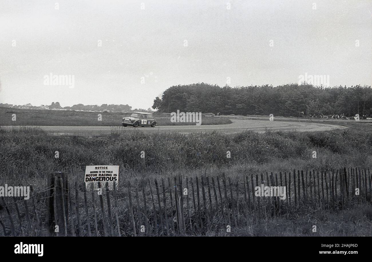 1960s, histórico, deporte del motor, un mini coche que compite alrededor de un circuito, Inglaterra, Reino Unido. Lejos de la pista, un pequeño aviso en la hierba por una cerca de madera corta dice, Motor Racing es PELIGROSO. Foto de stock