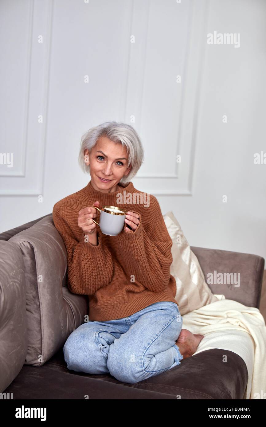 Mujer positiva con pelo gris corto en jeans y un cálido suéter sentado en un cómodo sofá y bebiendo una taza de café en una habitación luminosa Foto de stock
