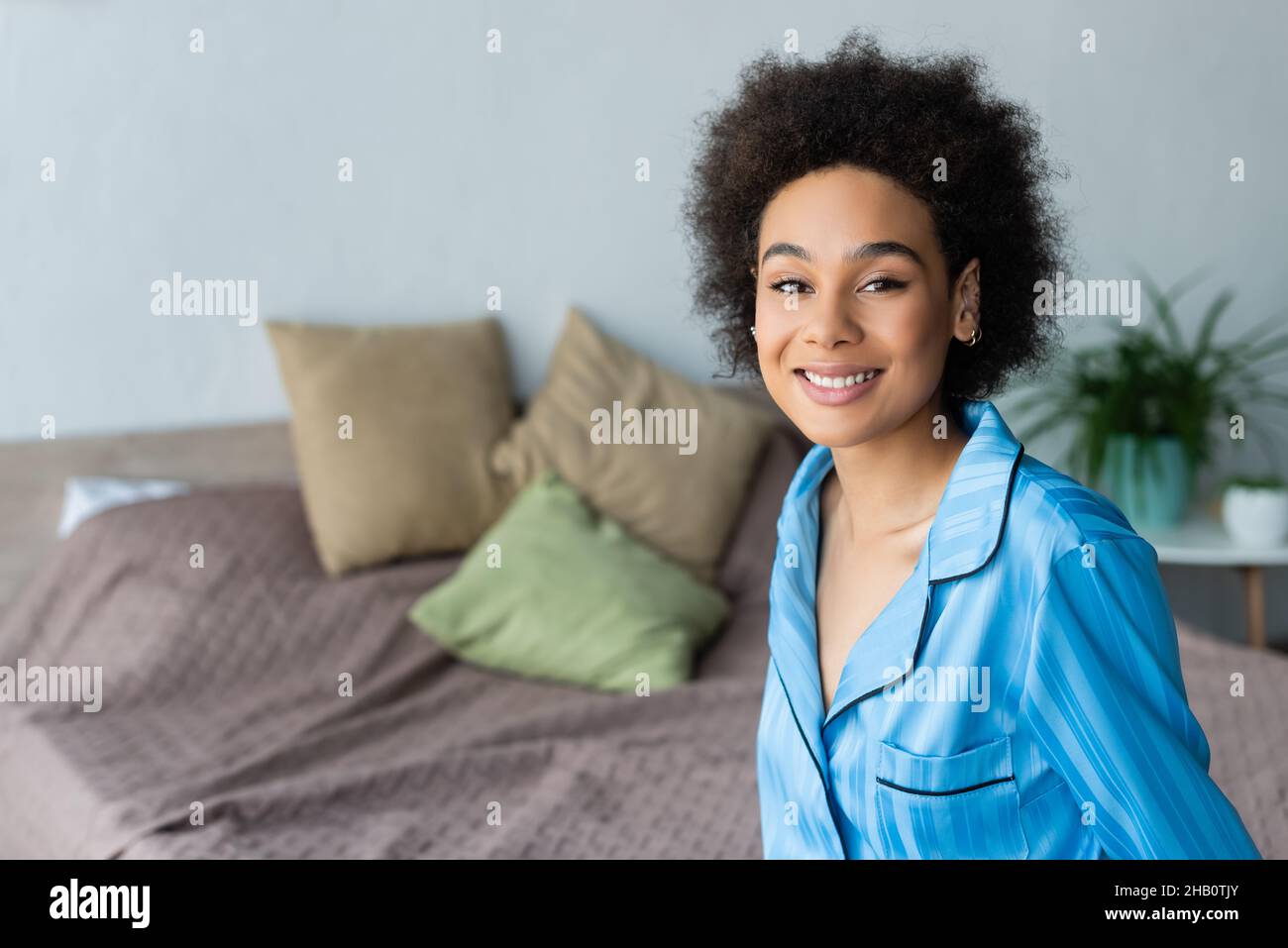 Mujer afroamericana positiva en pijama sonriendo con la cámara en el dormitorio Foto de stock