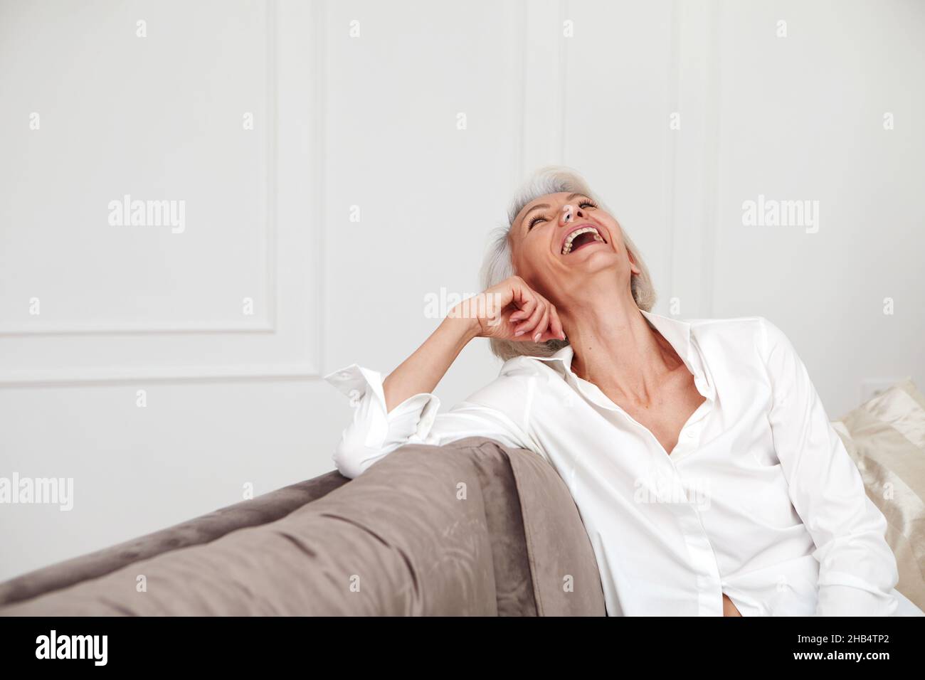 Contenido de la mujer de mediana edad en camisa blanca y con el pelo gris riendo mientras se sienta en el sofá en la sala de estar y divertirse Foto de stock