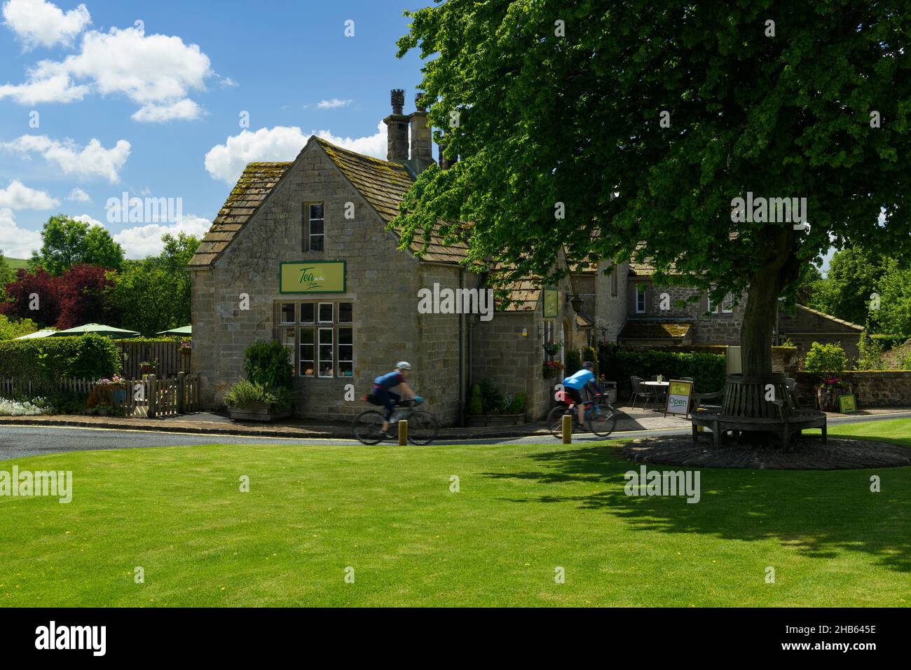 2 ciclistas pasando por pintorescas y atractivas cabañas de té café y árbol en el verde del pueblo, en la soleada abadía rural Bolton - Yorkshire Dales, Inglaterra Reino Unido. Foto de stock