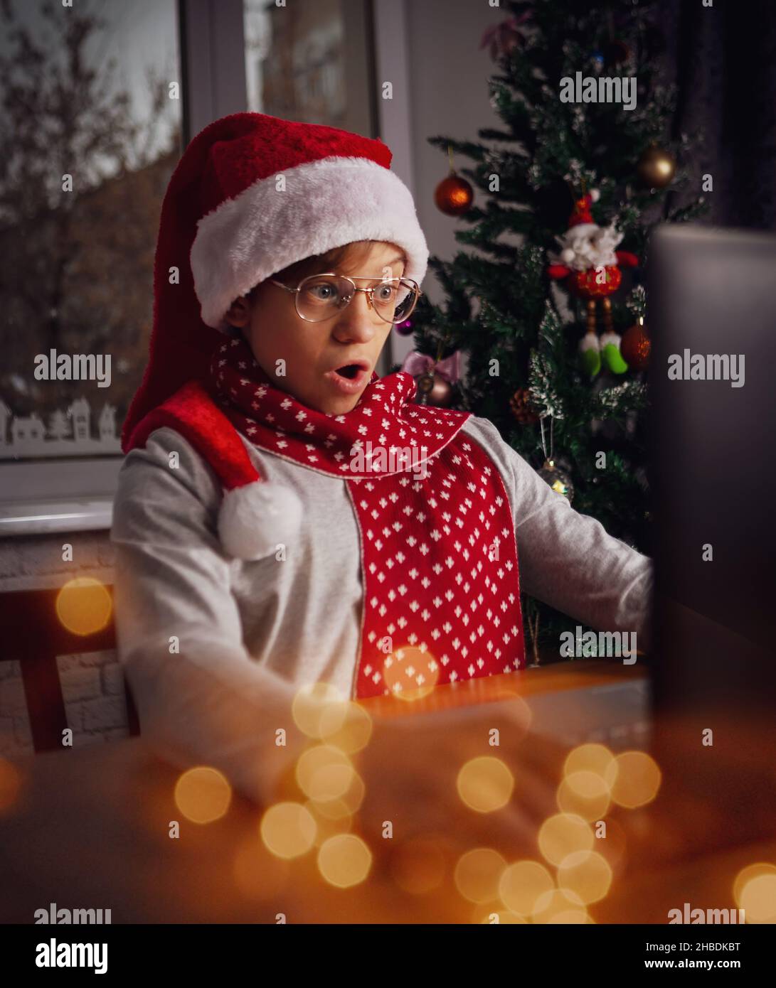 Divertido chico preguntándose sentado frente a la pantalla del portátil con gorra roja de Santa Claus teniendo una reunión de Navidad con familiares o amigos. Luces festivas. Foto de stock