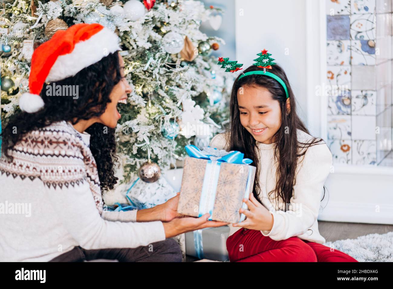 Rizado de pelo largo africano americano madre y la hija morena sentarse en la alfombra y abrir regalos festivos sonriendo feliz cerca del árbol de Navidad Foto de stock