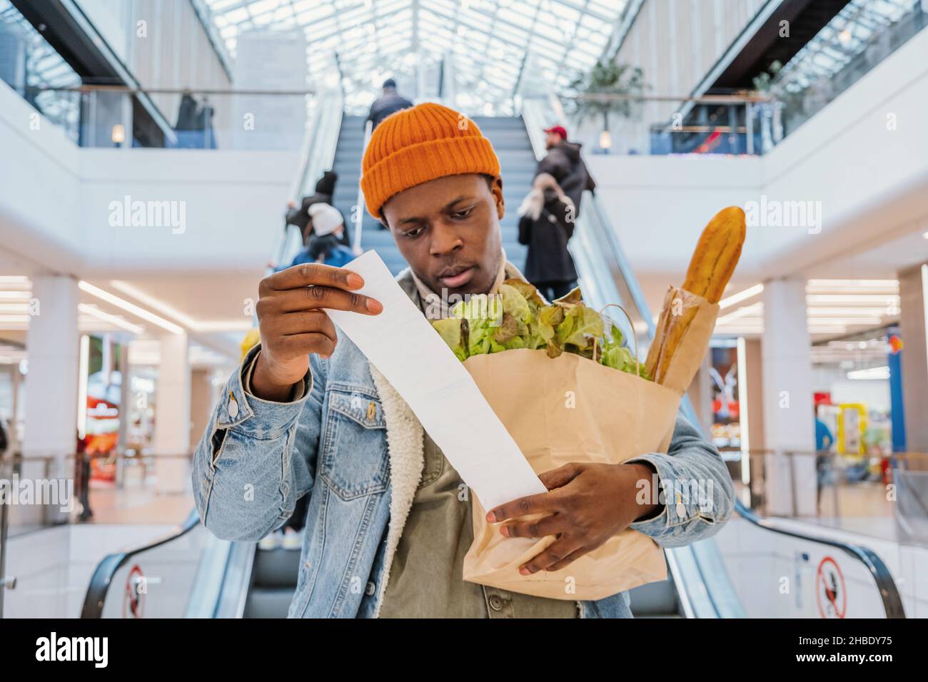 El hombre africano-americano sorprendido en la chaqueta del denim mira en el total del recibo en cheque de las ventas que sostiene la bolsa de papel con los productos en el centro comercial Foto de stock