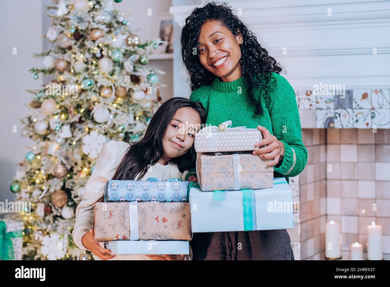 Alegre madre afroamericana e hija morena con pelo largo posan contra decoraciones de árbol de Navidad sosteniendo regalos Foto de stock