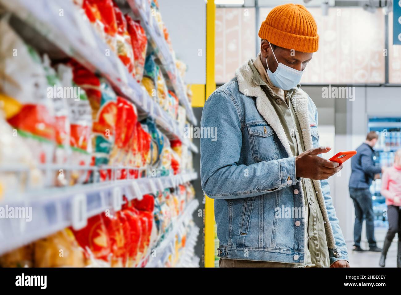 Un hombre hipster africano con una máscara médica y un sombrero de moda en un supermercado mira una lista de productos en su smartphone de pie en una estantería con productos y comprueba los productos para una cena festiva Foto de stock
