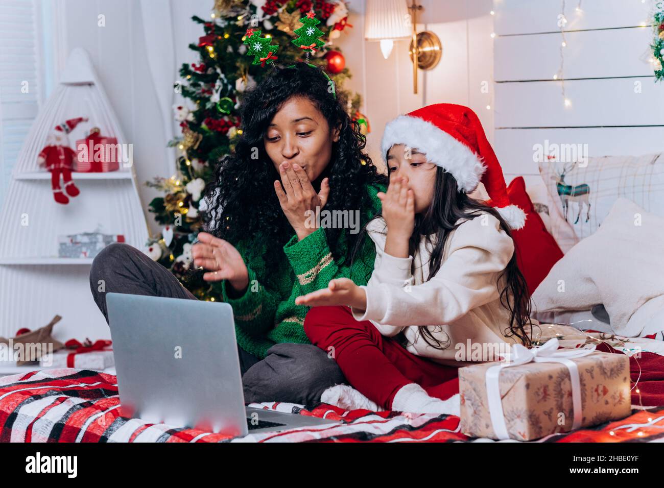 Una mujer afroamericana y una hija morena en el sombrero de Santa se sientan entre decoraciones navideñas y besos soplados charlando en videollamada a través de un ordenador portátil Foto de stock
