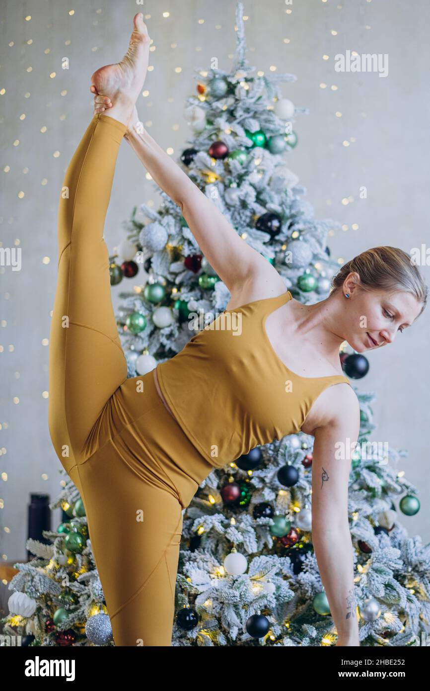 Mujer atlética con pelo rubio en traje deportivo de color amarillo que se extiende vertical contra el árbol de Navidad decorado con ornamentos y guirnaldas Foto de stock