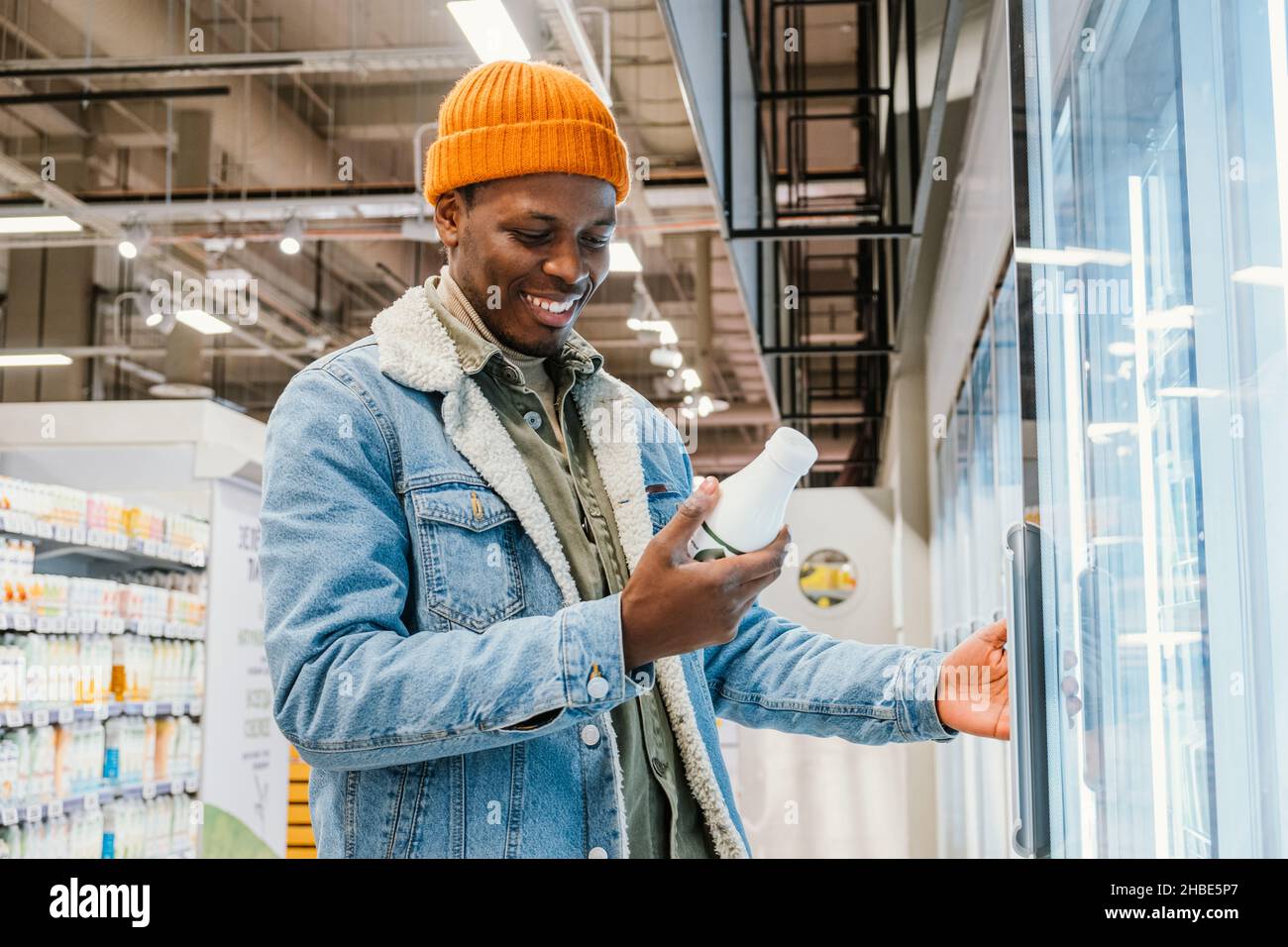 El hombre del milenio africano satisfecho en un sombrero de naranja en un supermercado elige la leche embotellada de pie en el refrigerador, y lee la composición del producto, sonrisas encontró el producto derecho Foto de stock