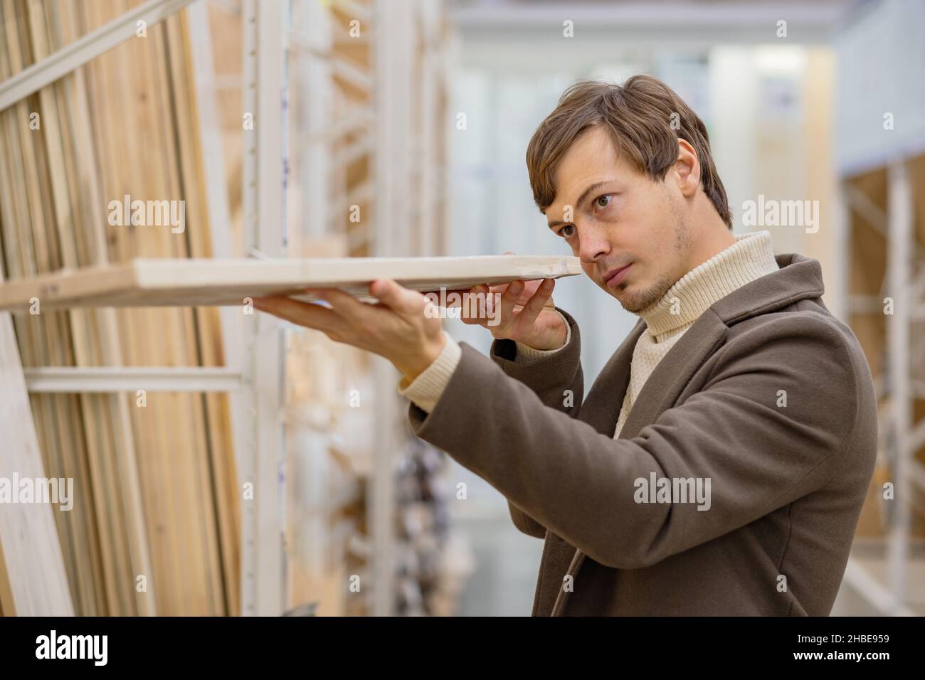 Un joven con pelo corto moreno con suéter y abrigo de abrigo elige una tabla de madera para la renovación del apartamento en el supermercado Foto de stock