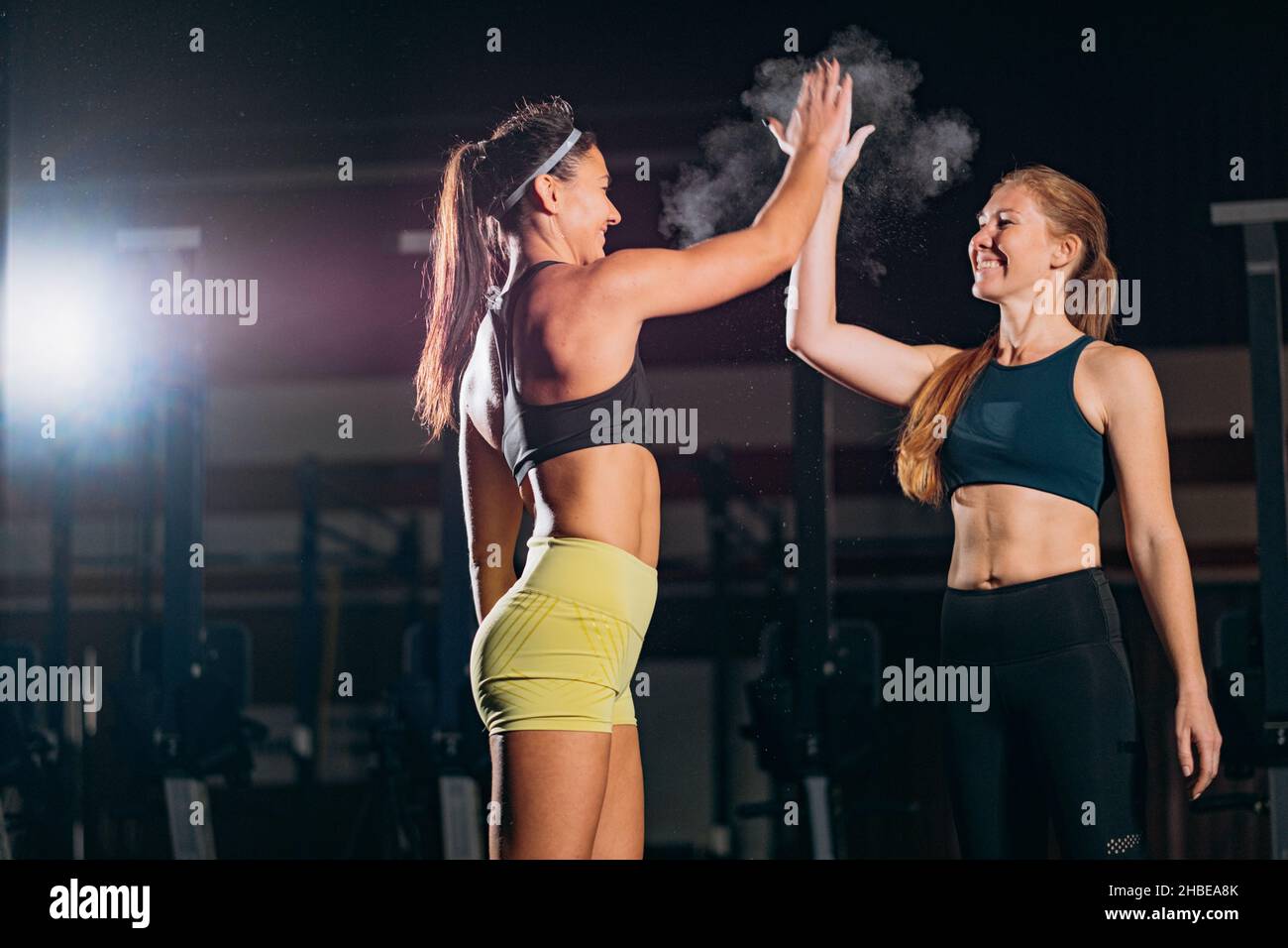 Las amigas que llevan trajes deportivos dan cinco alto a hacer ejercicio juntos y tomar pesas en el gimnasio contra la iluminación artificial Foto de stock