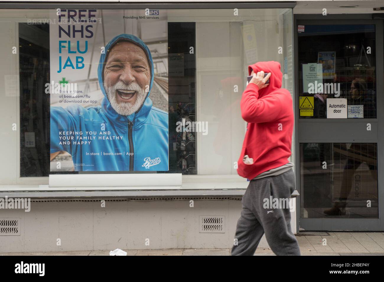 Hombre en el teléfono móvil en la sudadera roja caminar más allá de un cartel para la gripe de invierno jab, libre para los grupos elegibles de la gente Bexley Londres Inglaterra Foto de stock