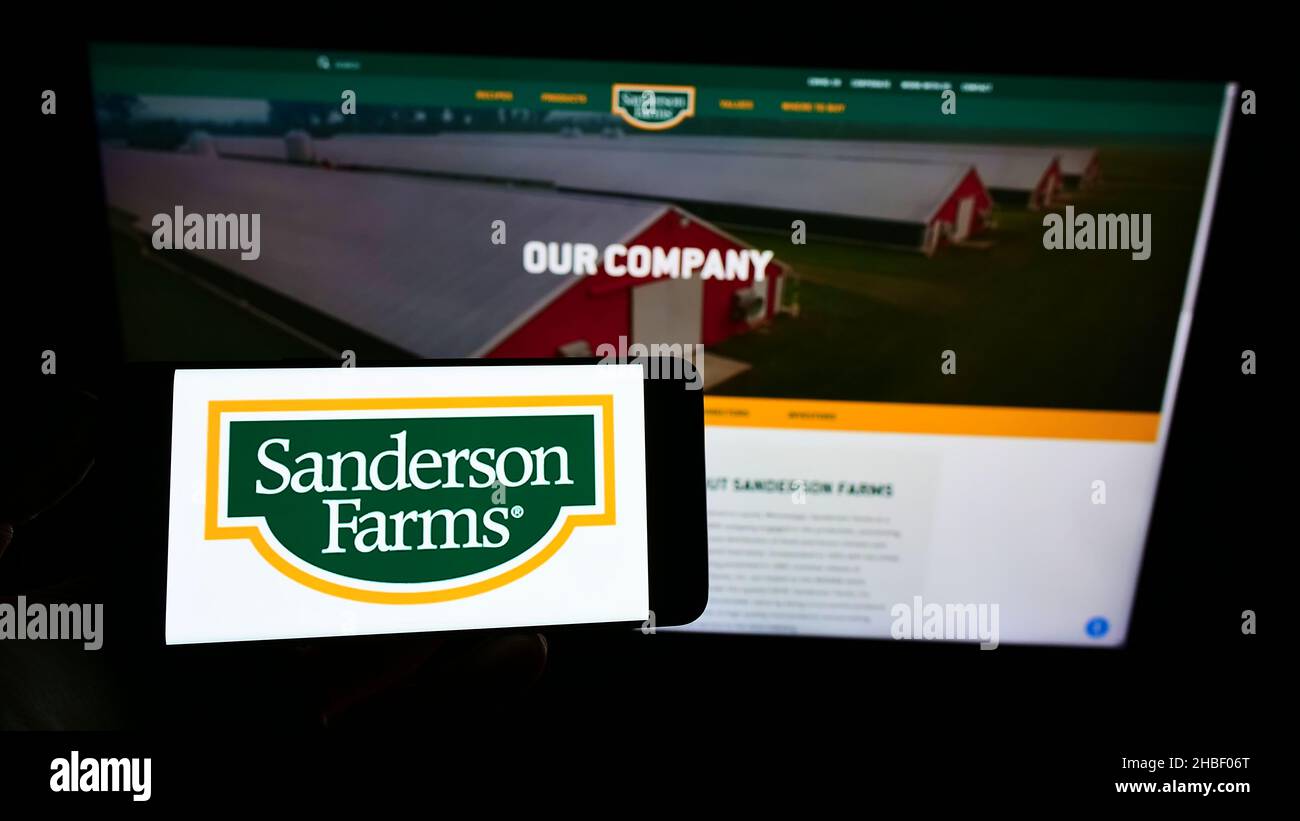Persona que sostiene el smartphone con el logotipo de la compañía avícola estadounidense Sanderson Farms Inc. En la pantalla frente al sitio web. Enfoque en la pantalla del teléfono. Foto de stock