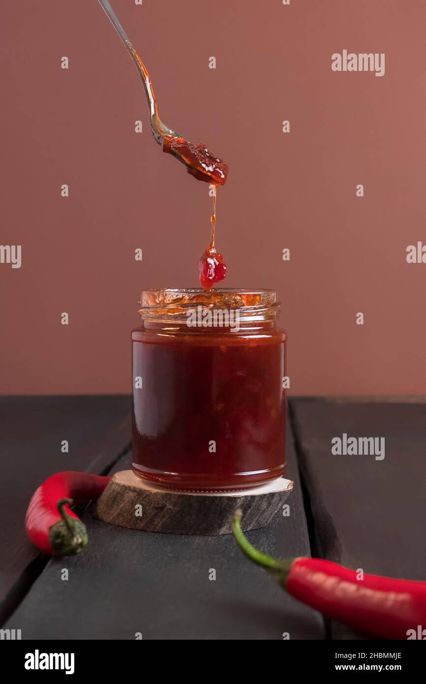 Cuchara con mermelada picante de pimiento rojo caliente cayendo en la jarra de mermelada sobre mesa de madera, enfoque selectivo Foto de stock