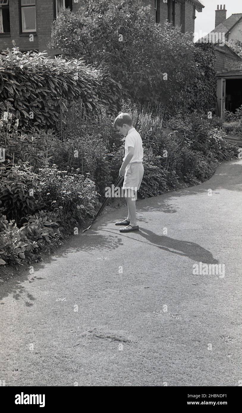 1950s, histórico, en un día soleado fuera en un jardín, un niño en pantalones cortos, camisa de manga corta y sandalias, ayudando con la jardinería, recortando el borde de un césped, utilizando un par de largos tirados o secateurs, Inglaterra, Reino Unido. Foto de stock