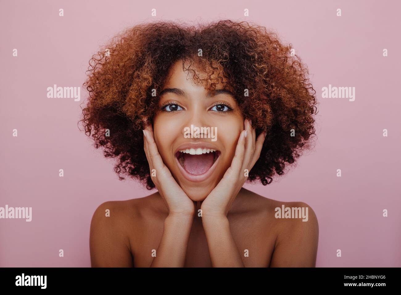 Retrato de una joven mujer de piel oscura agradablemente sorprendida con un cabello rizado exuberante mirando hacia la cámara con la boca abierta y su cara en sus manos sobre un fondo rosa sonriendo Foto de stock